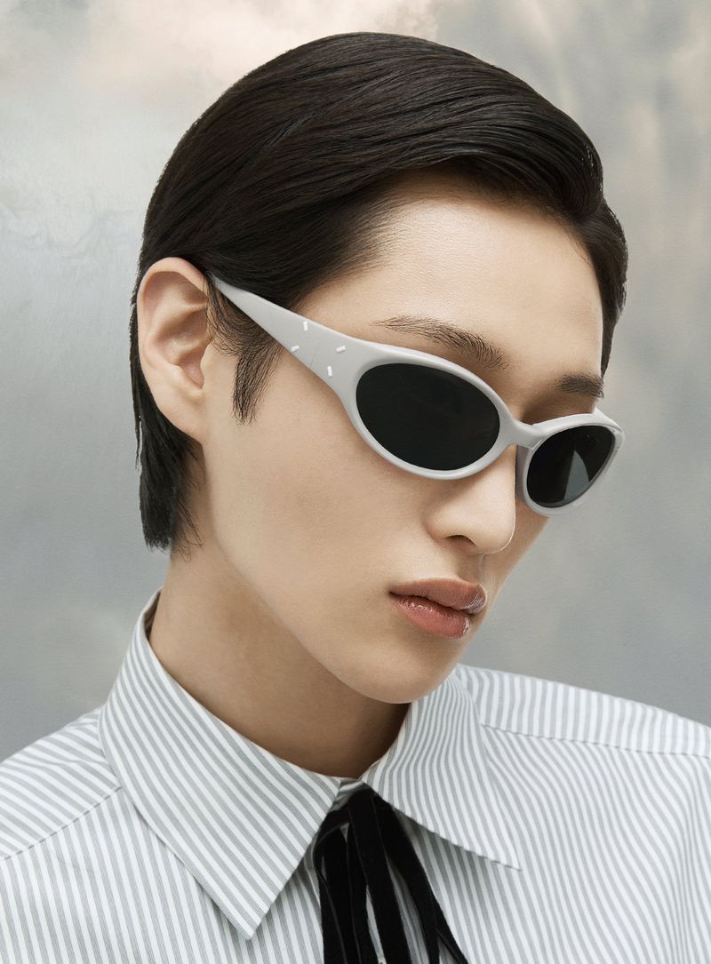 Close-up of a model wearing gray-framed sunglasses and a striped shirt with a black bow, set against a muted, cloudy background.