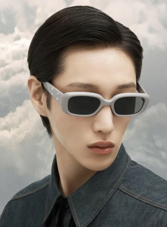 Portrait of a person wearing gray sunglasses and a denim jacket, posing against a soft cloud background, showcasing fashion eyewear.