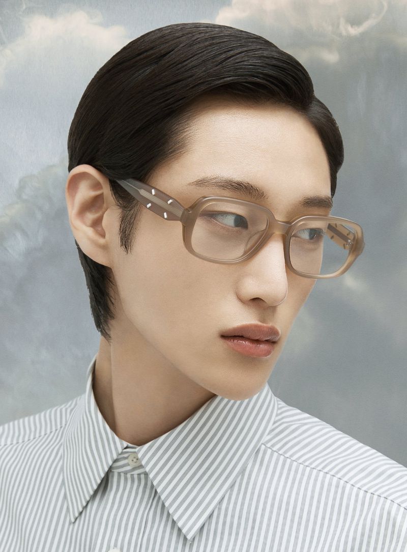 Close-up of an adult wearing square framed eyeglasses and a striped shirt, posing against a blurred cloud background in a studio.