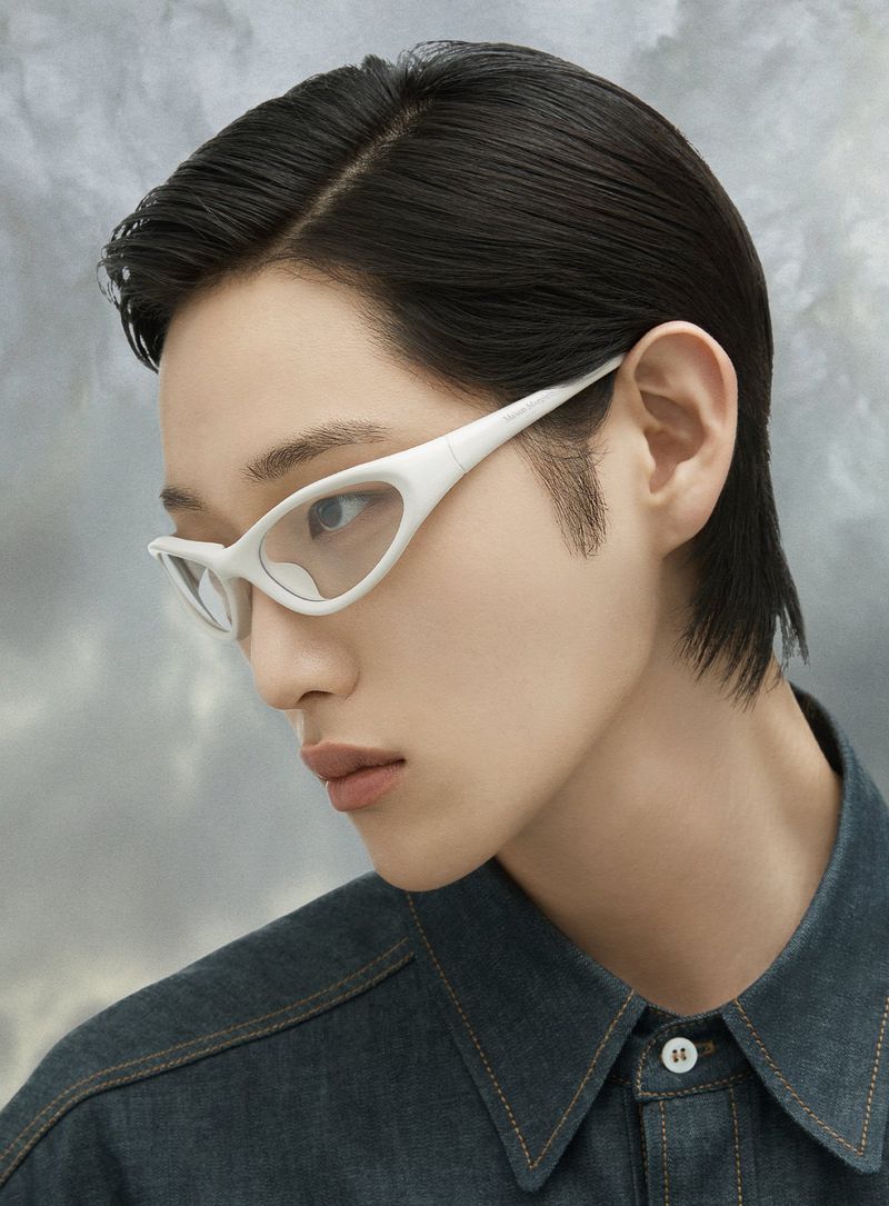 Portrait of a young adult with dark hair, wearing white-framed glasses and a dark denim shirt, set against a hazy cloudy backdrop.