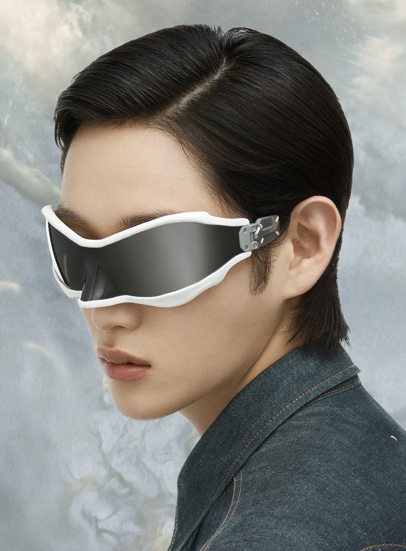 Close-up of a person wearing futuristic white rimmed visor-like sunglasses, denim jacket with a cloud-like background and soft lighting.