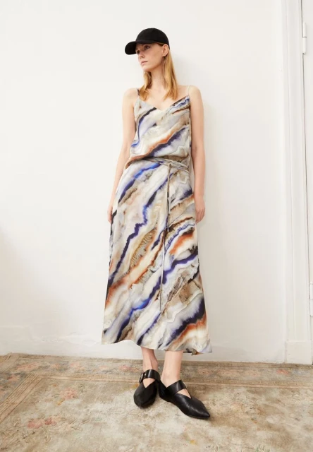 Fashion shot of a woman in a patterned top and skirt, wearing a baseball cap and shoes, posing against a white wall indoors.