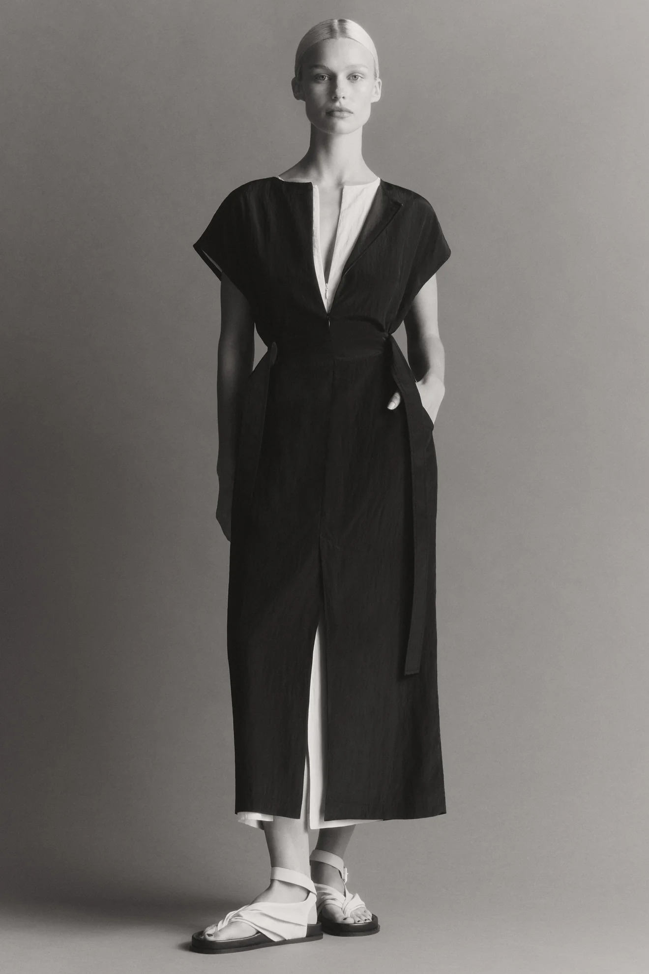 Full-length portrait of a fair-skinned woman wearing a black dress with a white inner layer and white sandals against a grey backdrop.