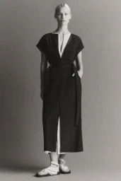 Model standing in a long dark dress and sandals against a plain grey backdrop for the A.Emery Resort Campaign 25.