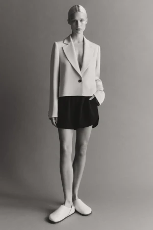Model standing against a neutral studio backdrop wearing a cropped cream blazer, dark shorts, and white shoes.
