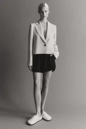 Model standing against a neutral studio backdrop wearing a cropped cream blazer, dark shorts, and white shoes.