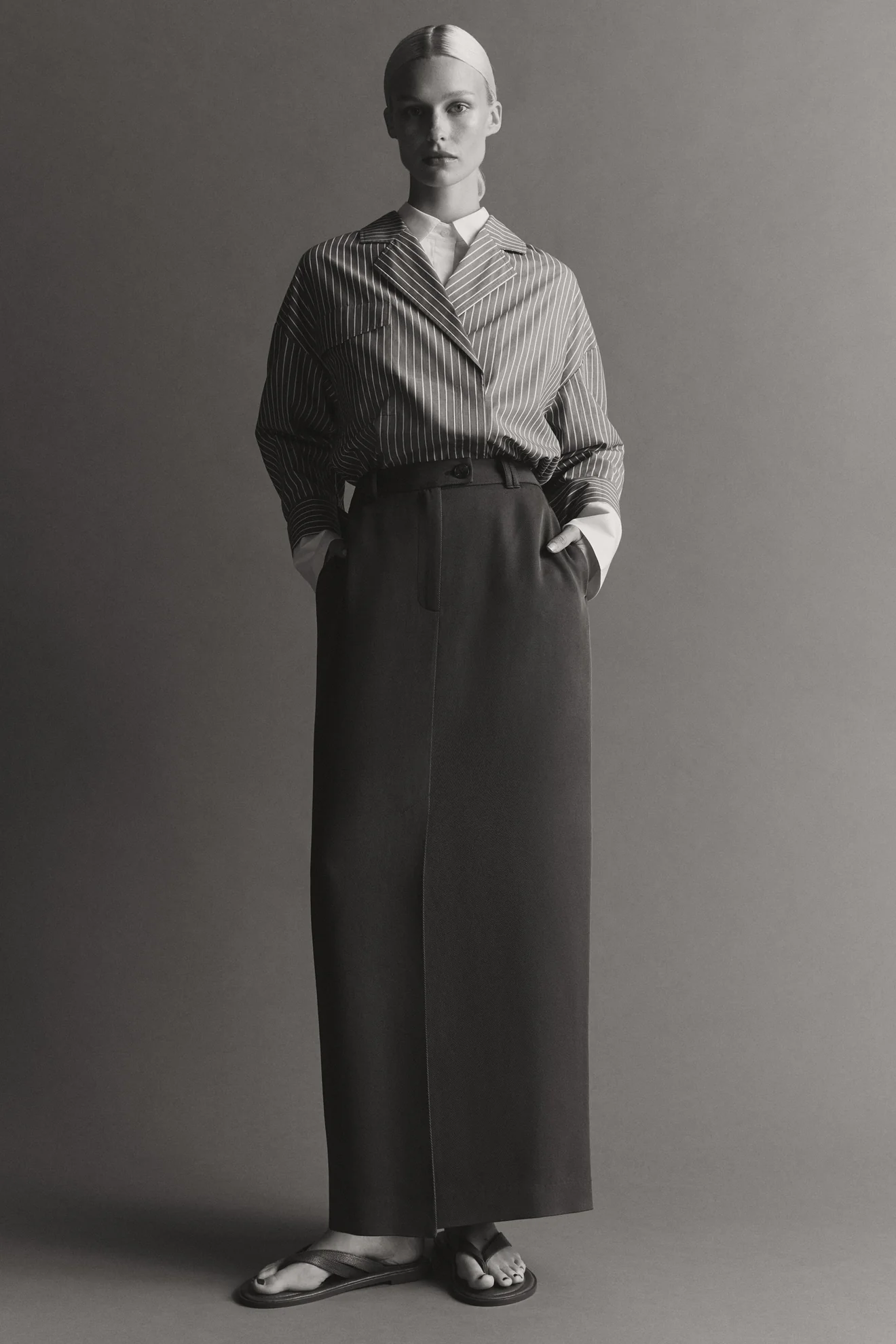 Model standing in a striped collared shirt and long dark maxi skirt against a solid grey background