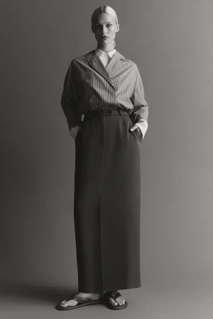 Model standing in a striped collared shirt and long dark maxi skirt against a solid grey background