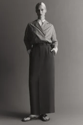 Model standing in a striped collared shirt and long dark maxi skirt against a solid grey background