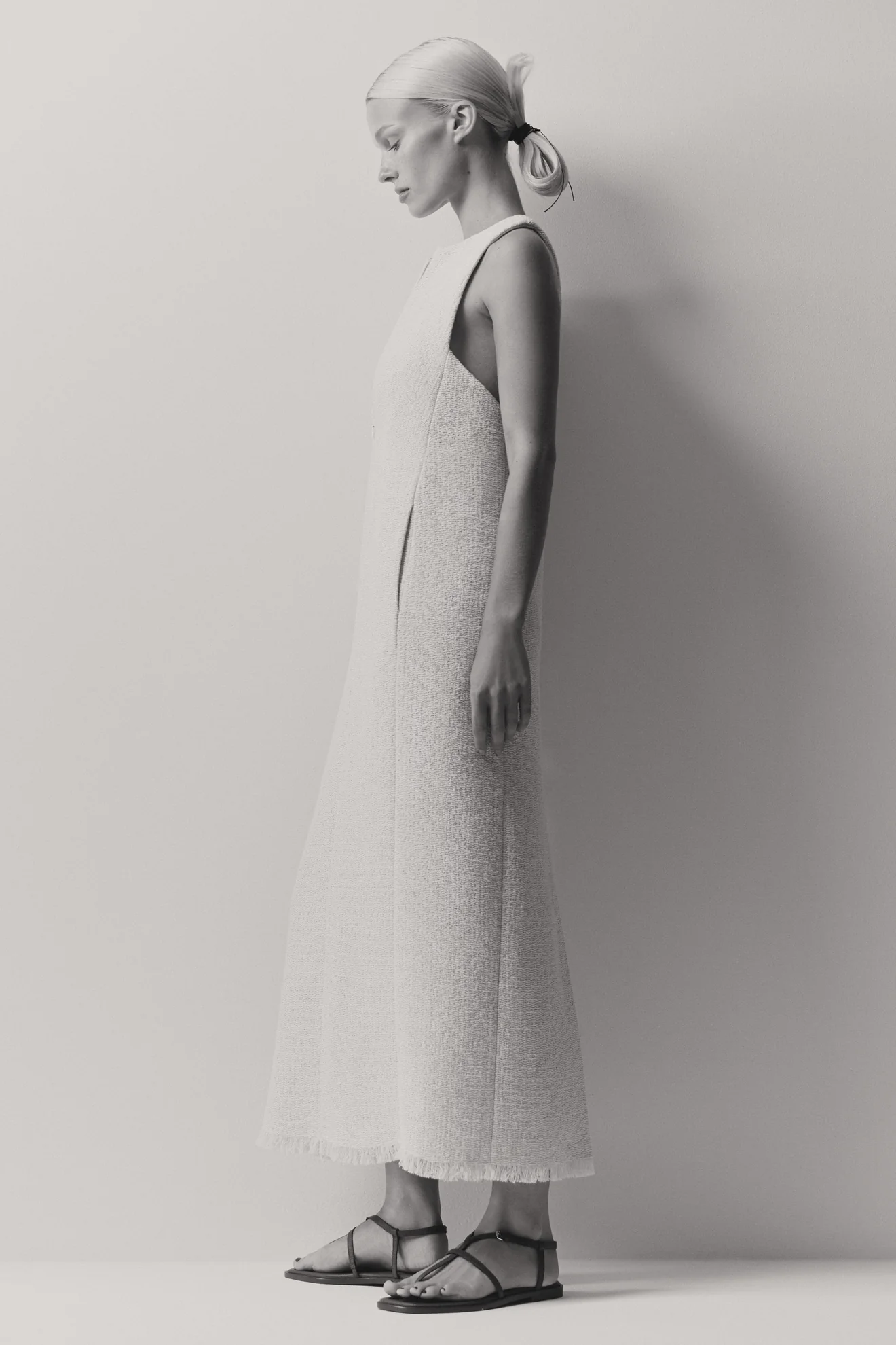 Model in profile wearing a textured cream dress and strappy flat sandals against a light grey studio background.