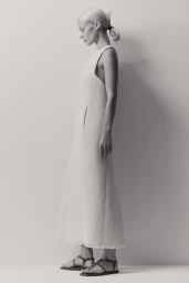 Model in profile wearing a textured cream dress and strappy flat sandals against a light grey studio background.
