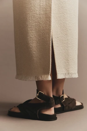 Close-up of a woman wearing a textured midi skirt with fringe and dark brown buckled sandals, set against a neutral backdrop.