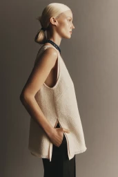 Model in profile wearing a cream sleeveless knit top and dark trousers against a neutral grey studio background