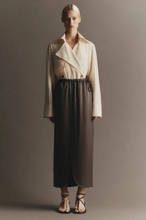 Fashion portrait of a fair-skinned adult woman in a beige jacket, brown skirt, and strappy sandals against a neutral background.
