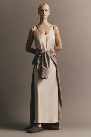 Model wearing cream silk slip dress and striped shirt tied around waist against a neutral backdrop