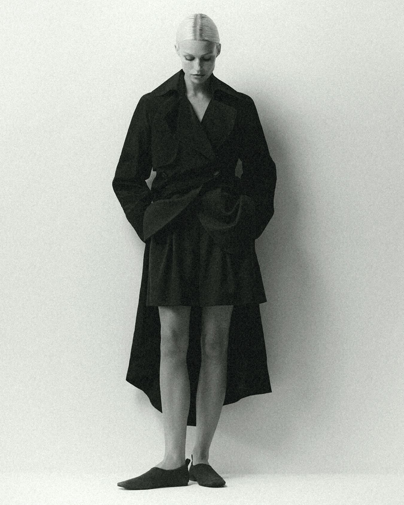 Monochrome fashion portrait of a slender woman in a dark coat and skirt looking down against a light background in a photo studio.