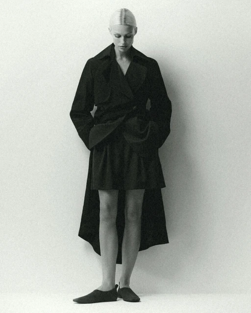 Monochrome fashion portrait of a slender woman in a dark coat and skirt looking down against a light background in a photo studio.