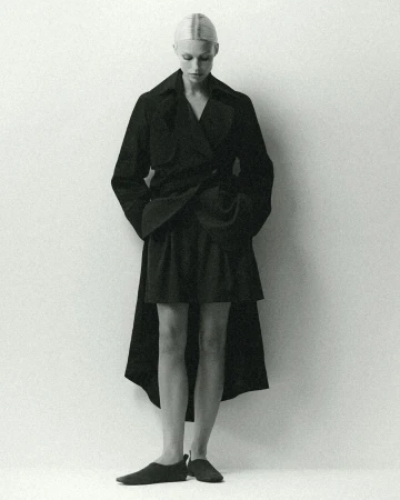 Monochrome fashion portrait of a slender woman in a dark coat and skirt looking down against a light background in a photo studio.