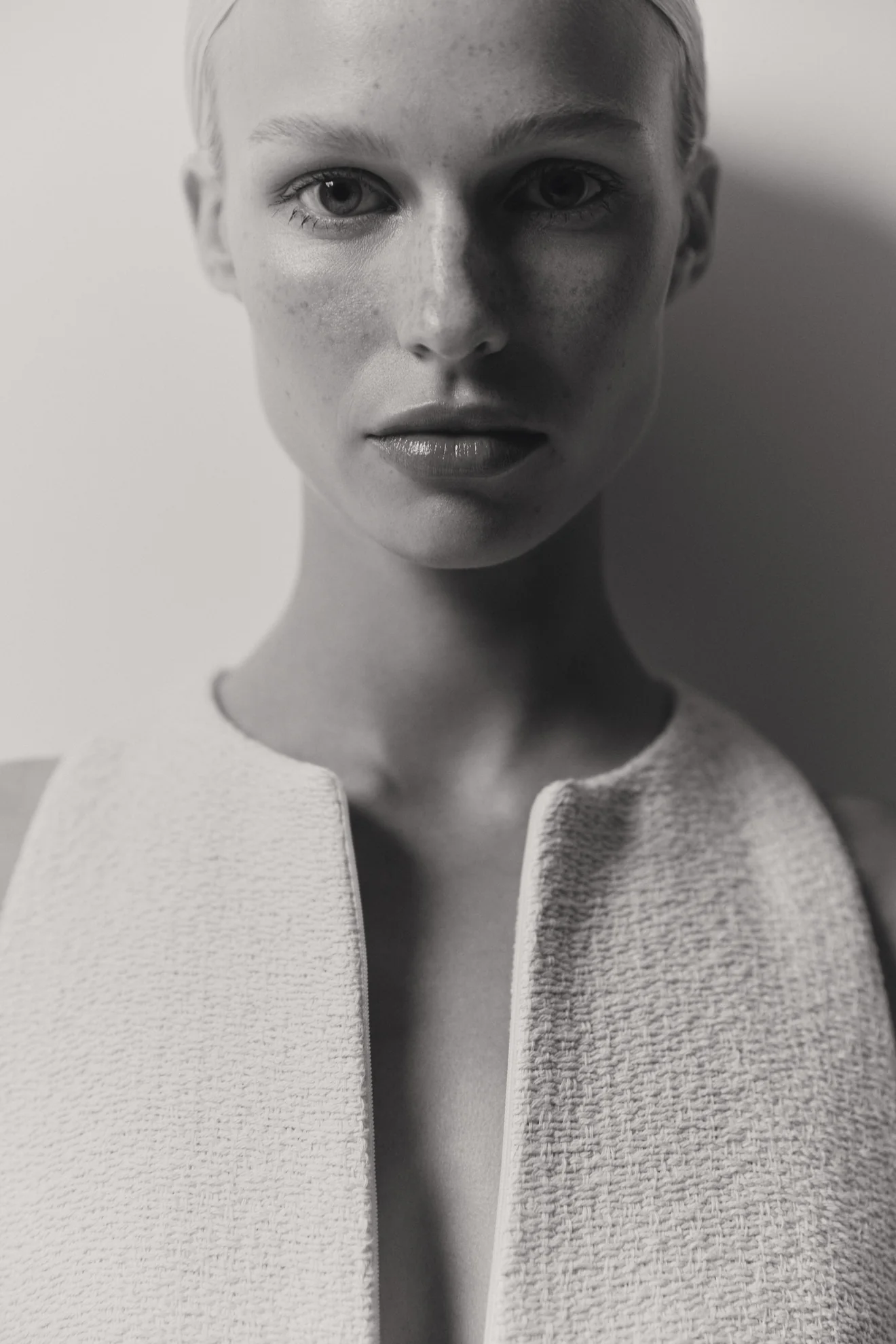Close-up portrait of a female model wearing a textured white sleeveless top against a plain light wall.