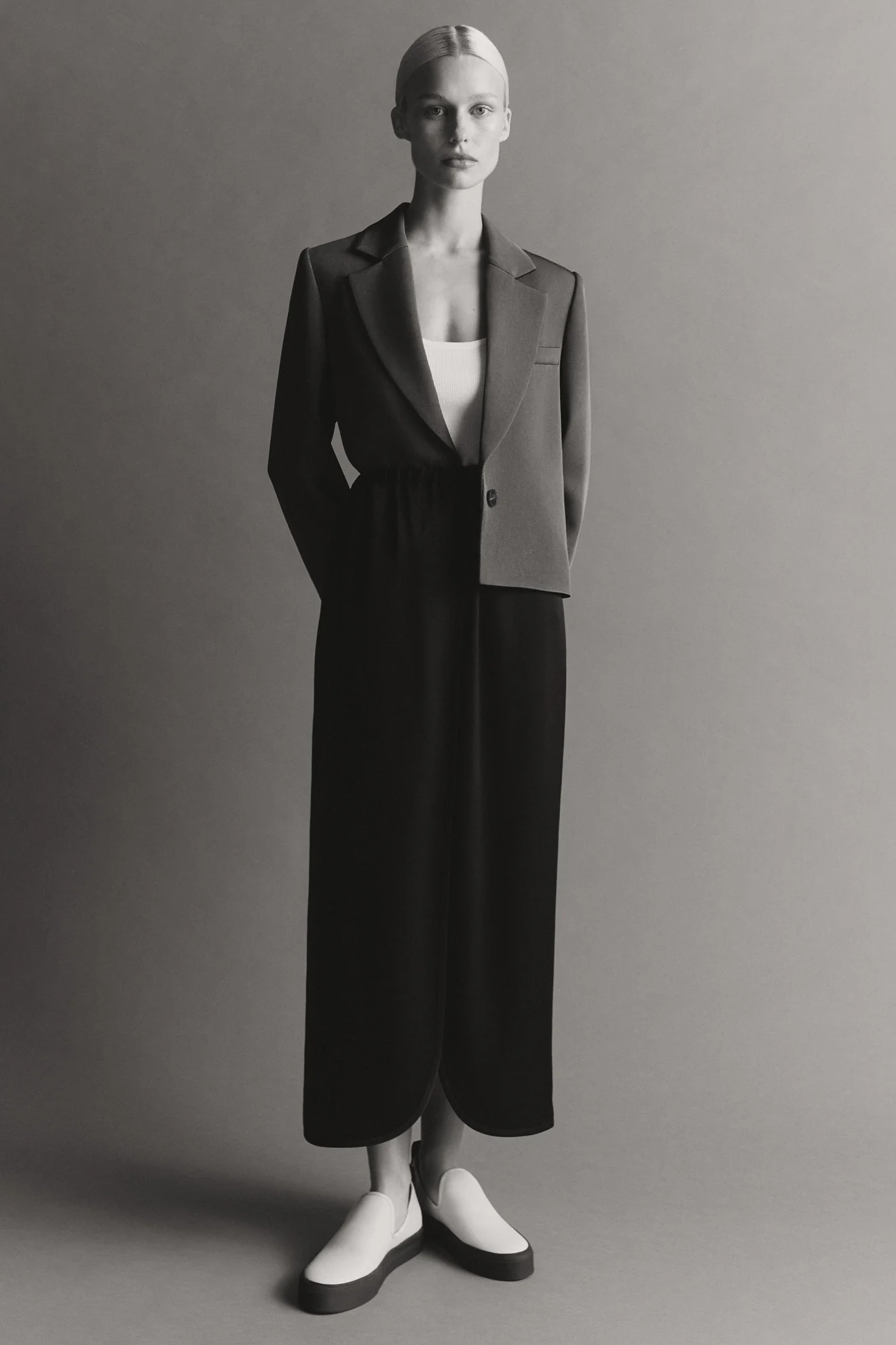 Model standing against a neutral studio backdrop wearing an unstructured blazer, dark skirt, and light slip-on shoes.