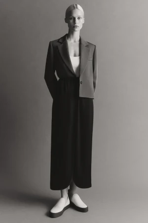 Model standing against a neutral studio backdrop wearing an unstructured blazer, dark skirt, and light slip-on shoes.