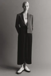 Model standing against a neutral studio backdrop wearing an unstructured blazer, dark skirt, and light slip-on shoes.