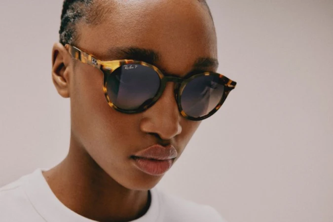 Close-up of an adult woman with dark skin and short hair, wearing patterned sunglasses, posing in front of a neutral background.