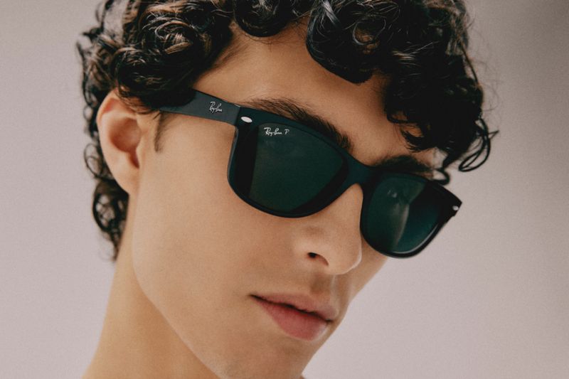 Close-up of a curly-haired adult male wearing black Ray-Ban sunglasses against a neutral backdrop, showcasing a fashion accessory.