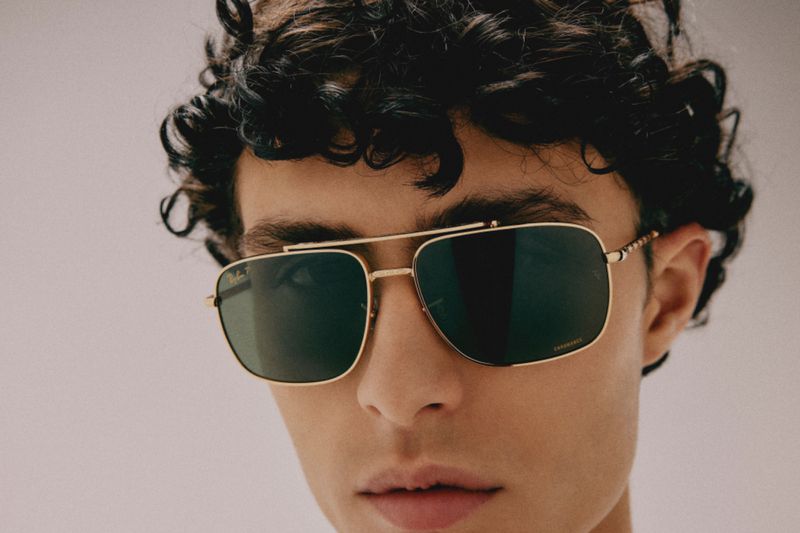 Close-up of adult male wearing aviator sunglasses with a gold frame. The man has curly dark hair, and the background is blurred, neutral tone.