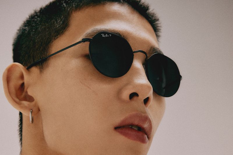 Close-up of an adult with an earring, wearing black Ray-Ban sunglasses. The background is out of focus, with a light neutral tone.