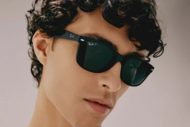 Close-up portrait of a young man wearing black sunglasses with dark curly hair against a neutral background