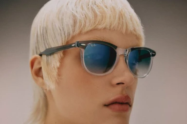 Close-up portrait of a model with blonde hair wearing dark framed sunglasses by Ray-Ban against a plain neutral background.