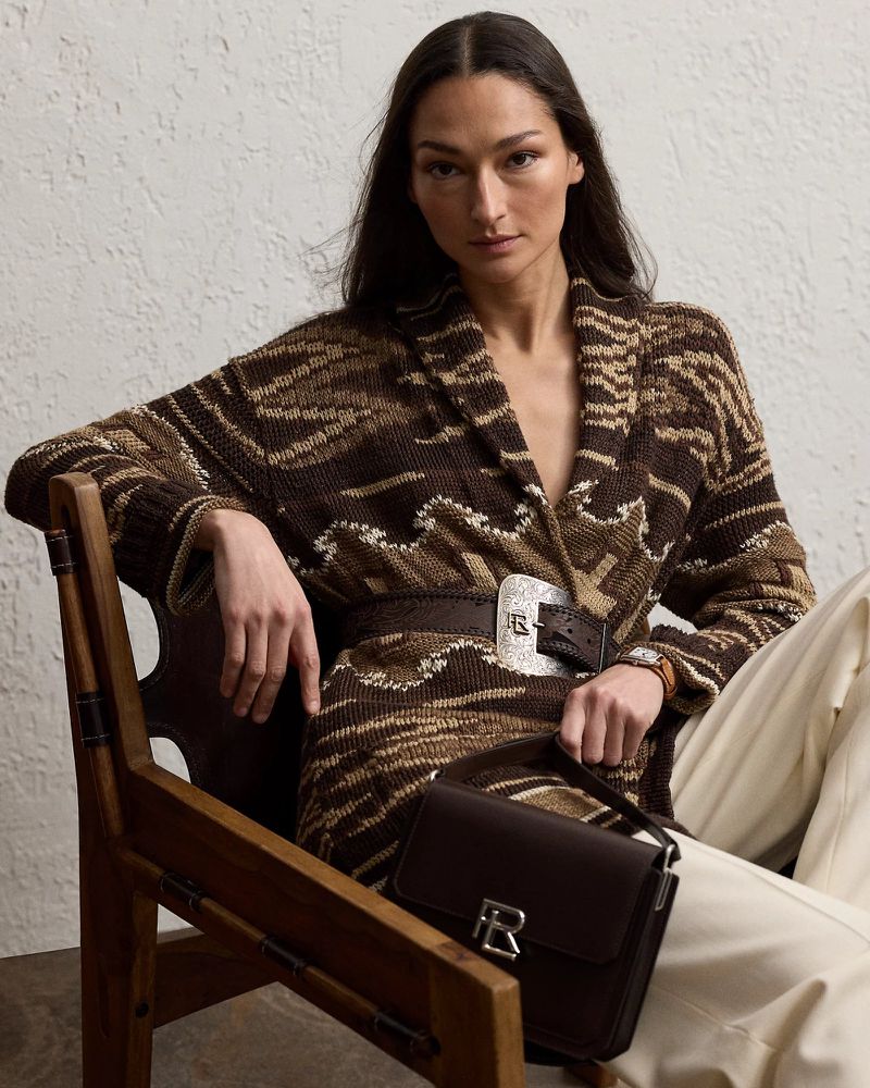 A woman in a patterned brown cardigan poses indoors, seated in a wood chair while holding a leather handbag in a neutral setting.
