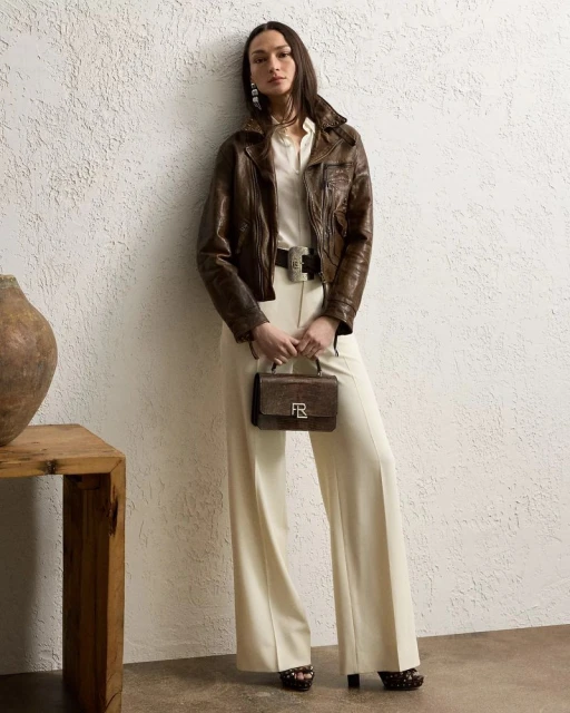 A slender adult female fashion model poses in an indoor photo studio wearing a brown leather jacket and cream-colored wide-leg trousers.