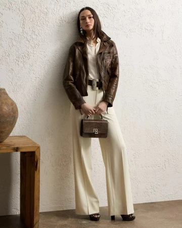 A slender adult female fashion model poses in an indoor photo studio wearing a brown leather jacket and cream-colored wide-leg trousers.