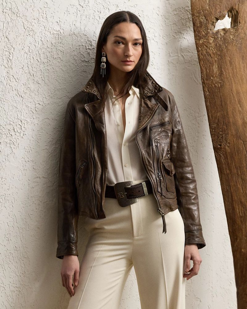 A woman models a dark brown leather jacket, cream blouse, and trousers indoors against a textured wall with rustic wood trim.