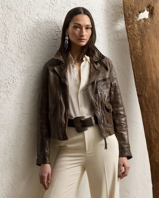 A woman models a dark brown leather jacket, cream blouse, and trousers indoors against a textured wall with rustic wood trim.