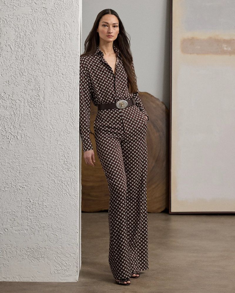 A brunette woman in a patterned brown and beige long-sleeved shirt and wide-legged pants, accessorized with a brown belt, poses indoors.
