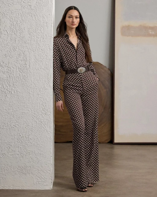 A brunette woman in a patterned brown and beige long-sleeved shirt and wide-legged pants, accessorized with a brown belt, poses indoors.