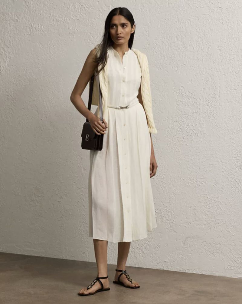 Elegant woman in a white sleeveless dress with a belt, paired with flat sandals and a brown purse in front of a textured wall.