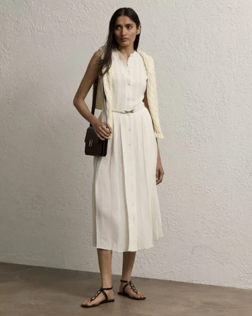Elegant woman in a white sleeveless dress with a belt, paired with flat sandals and a brown purse in front of a textured wall.