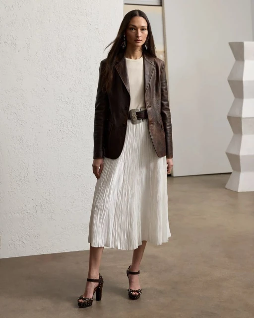 Elegant woman in a dark brown leather blazer, a white pleated skirt, and dark high heels strikes a pose in a minimalist studio.