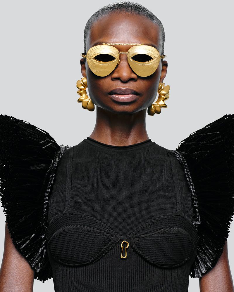 A close-up of a Black woman wearing gold sunglasses and large gold earrings, against a light gray background in a studio shot.