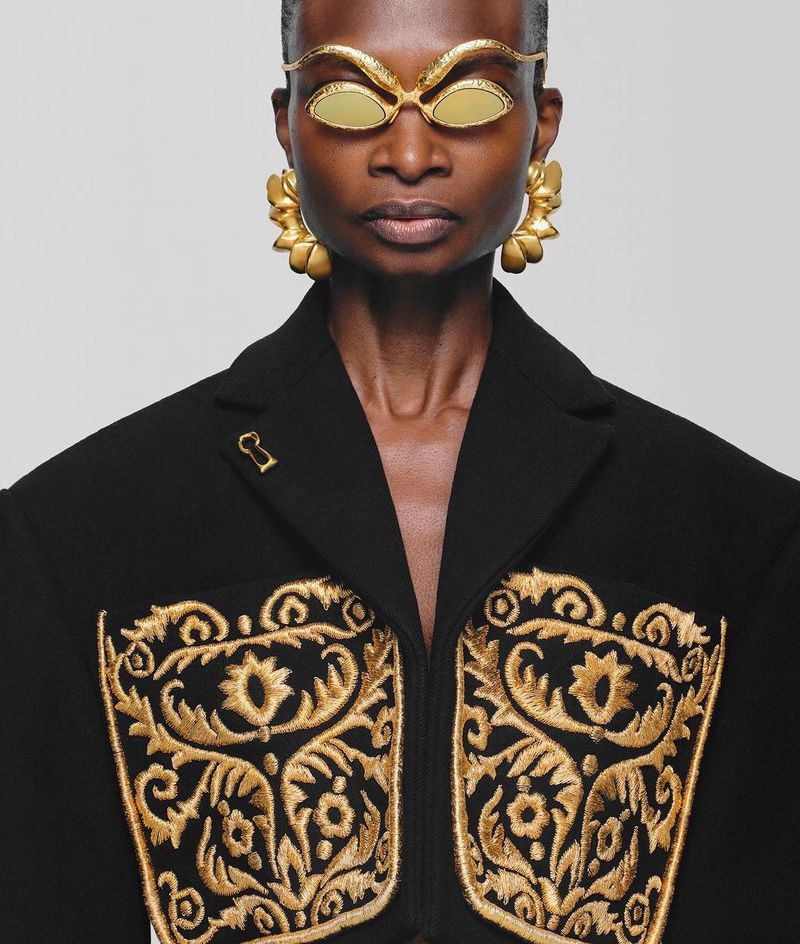 Eye-level shot of an adult woman with dark skin wearing gold sunglasses and gold floral earrings, adorned black coat with gold embroidery.