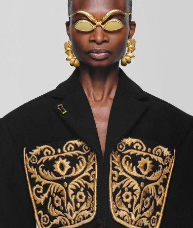 Eye-level shot of an adult woman with dark skin wearing gold sunglasses and gold floral earrings, adorned black coat with gold embroidery.