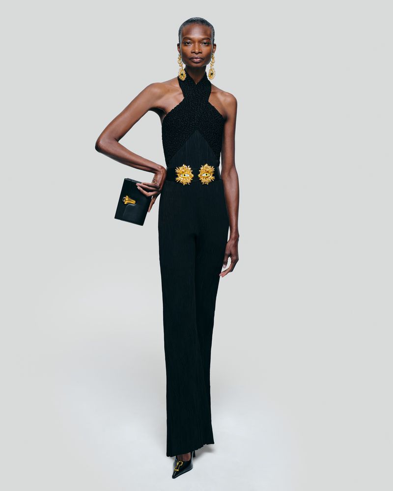 Elegant adult woman in black halter jumpsuit, statement earrings, and heels poses in a bright studio with a black clutch.