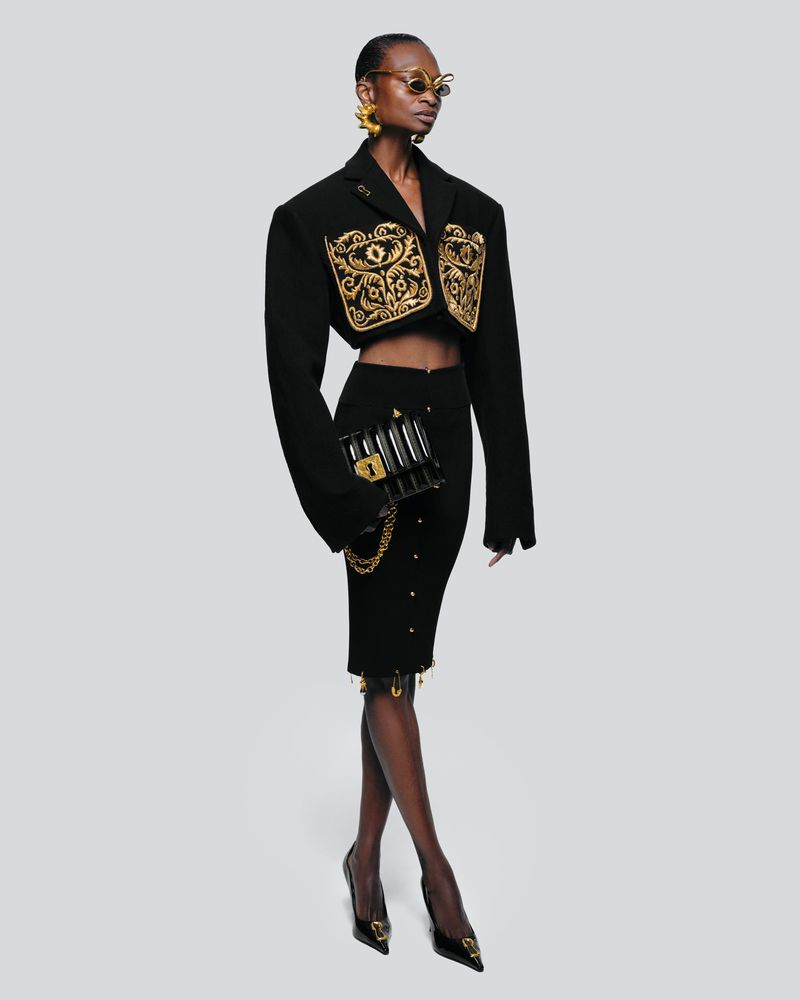 Full-length studio shot of a Black female model wearing a black blazer with gold embroidery, black skirt, black heels and holding a purse.