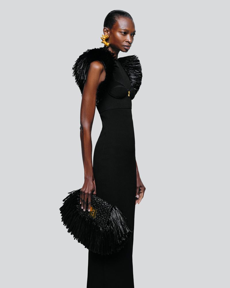 A striking fashion shot featuring a Black woman in an elegant black gown with feathered details, holding a fringed clutch, against a grey backdrop.