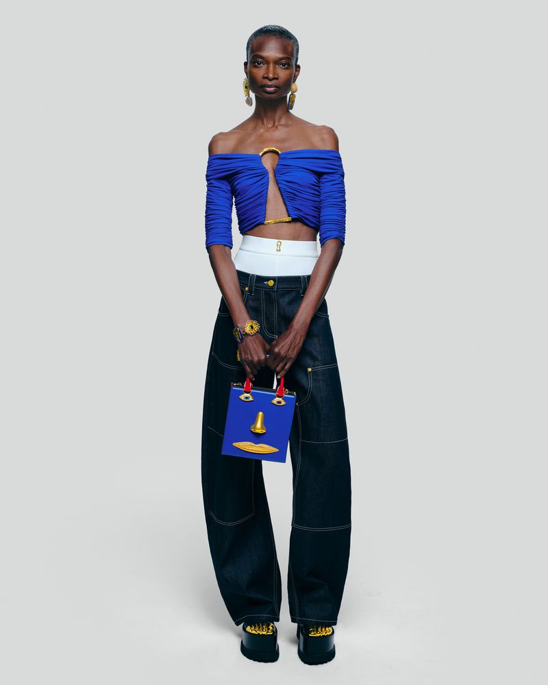 A woman stands in a studio wearing a blue top and baggy denim jeans, carrying a blue handbag with a yellow face design.