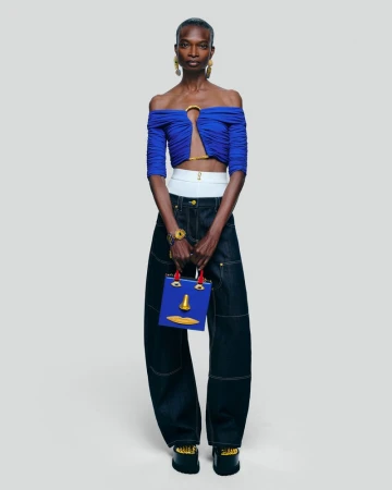 A woman stands in a studio wearing a blue top and baggy denim jeans, carrying a blue handbag with a yellow face design.